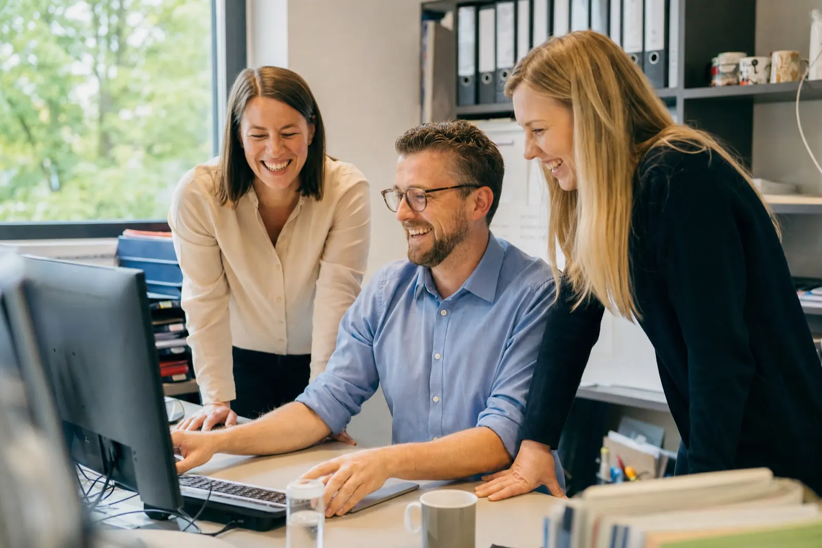 Drei Mitarbeitende in einem Büro schauen gemeinsam lachend auf einen Bildschirm